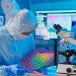 asian male technician in sterile coverall holds wafer that reflects many different colors with gloves and check it at semiconductor manufacturing plant