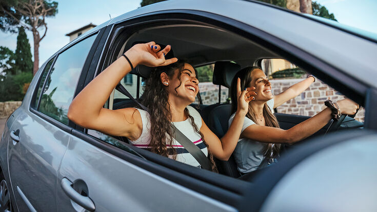 Two young women sing a song on the radio and dance in the car on a day trip in the summer - Best friends having fun together driving around the countryside - Smiling millennial in a relaxing moment